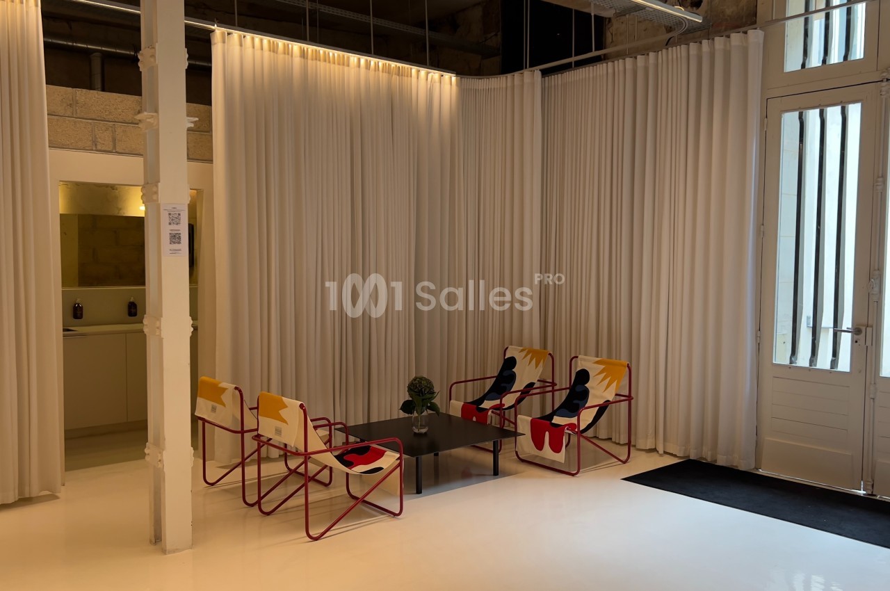 Espace lumineux avec des chaises colorées autour d'une table basse, entouré de rideaux blancs et d'une porte vitrée.