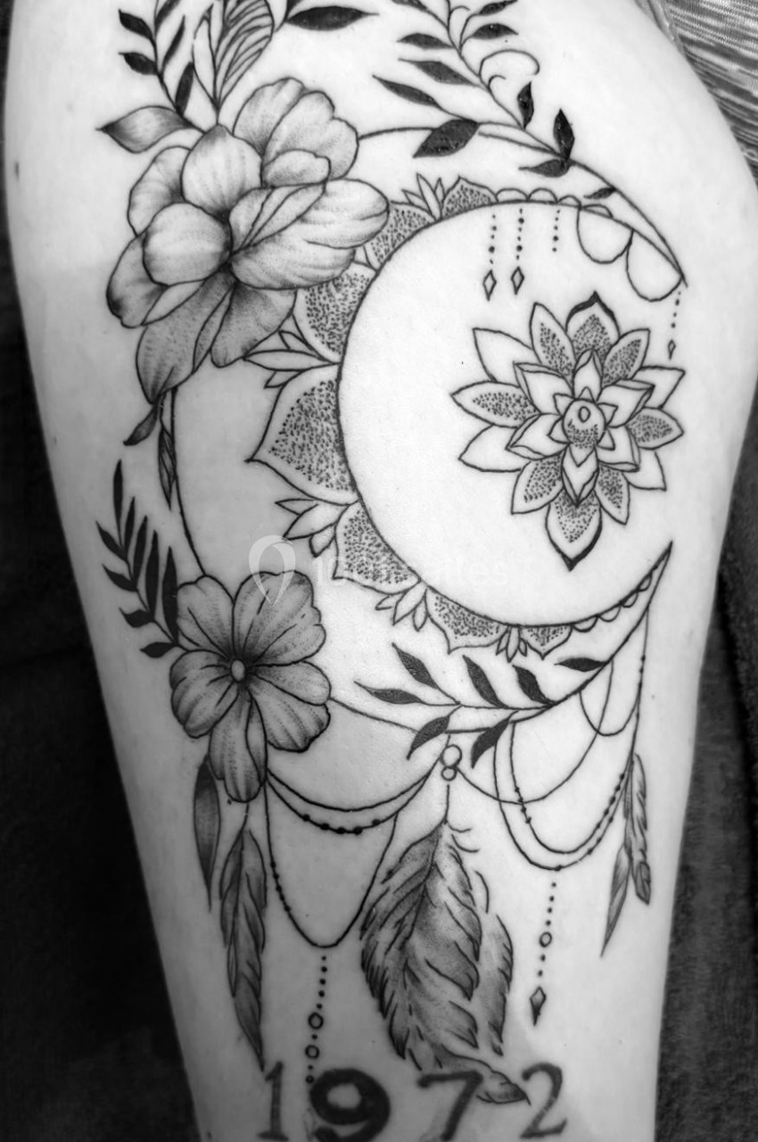 Tatouage représentant une lune ornée de fleurs, plumes, motifs géométriques et l'année ’1972’ en bas.