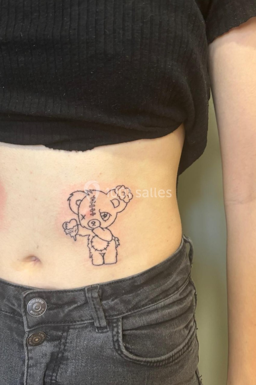 Tatouage d'un ours en peluche avec des détails de couture, situé sur le côté de l'abdomen d'une personne.