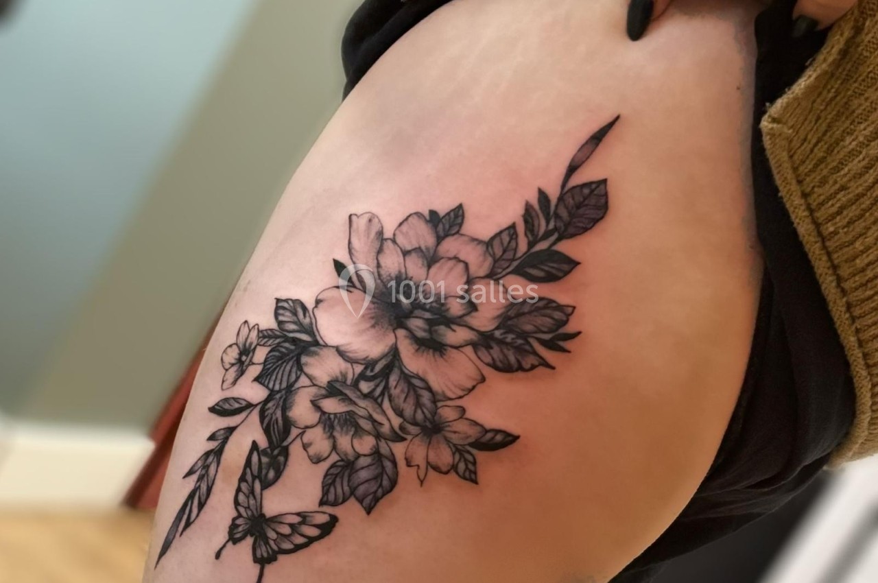 Tatouage floral noir et gris sur une cuisse, représentant des fleurs et des feuilles avec un papillon.