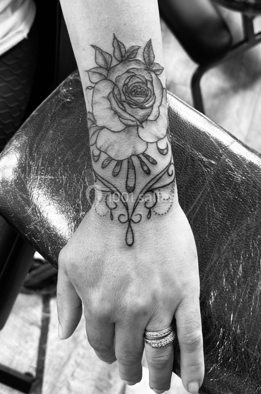 Tatouage de rose détaillé avec motifs ornementaux sur l'avant-bras d'une personne, appuyée sur un accoudoir.