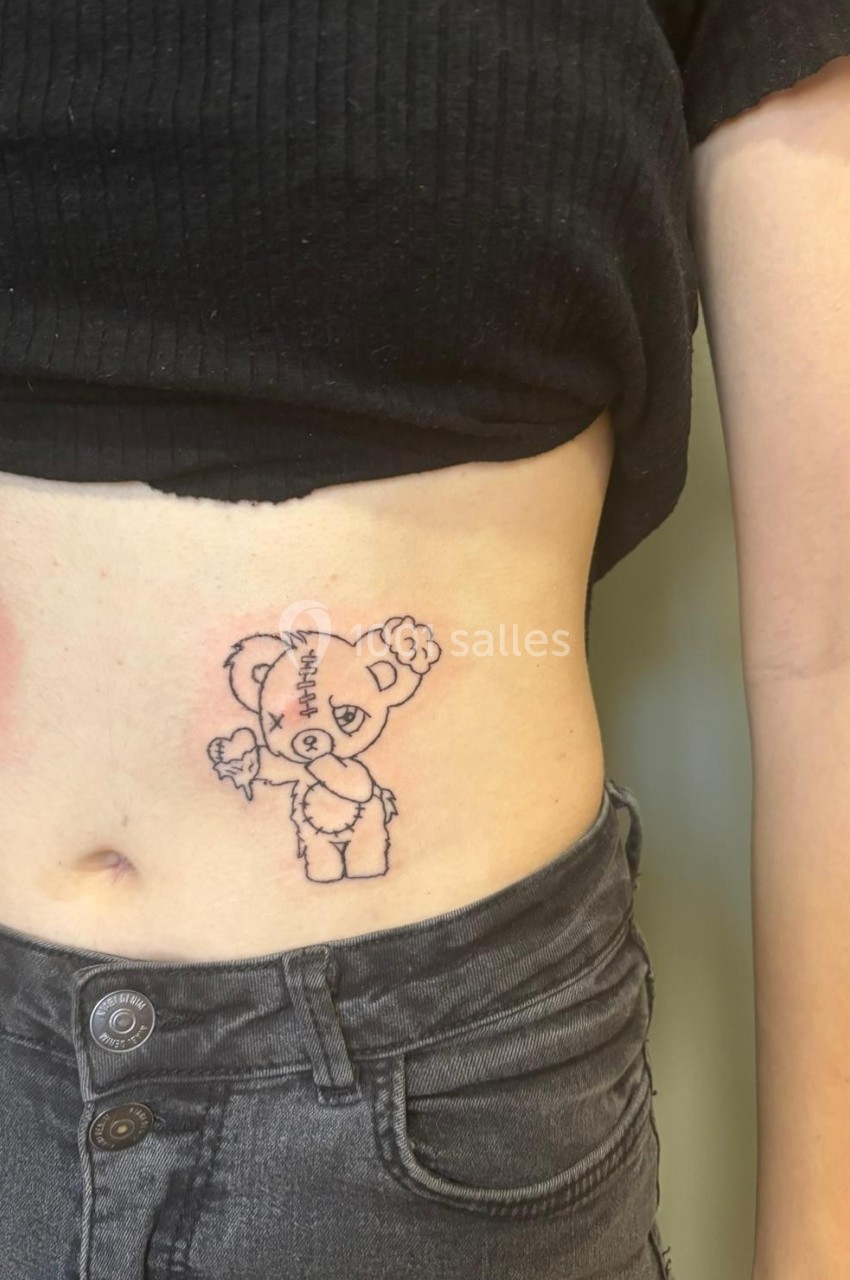 Tatouage d'un ours en peluche avec des coutures sur le ventre d'une personne portant un haut noir et un jean gris.