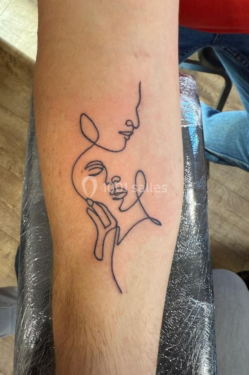 Tatouage minimaliste sur l'avant-bras représentant deux visages entrelacés dessinés en un seul trait.
