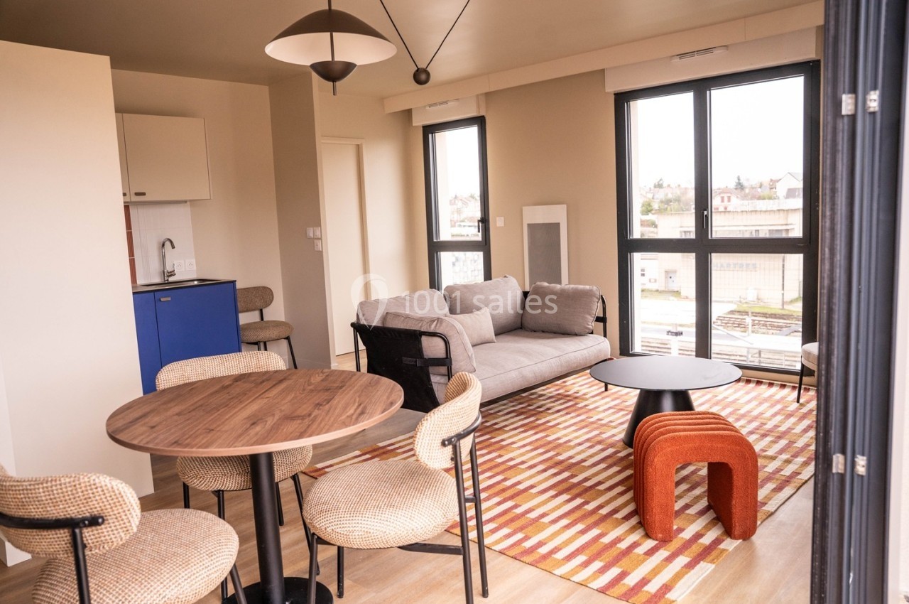 Salon lumineux avec canapé gris, table ronde en bois, chaises, tapis coloré et kitchenette en arrière-plan.