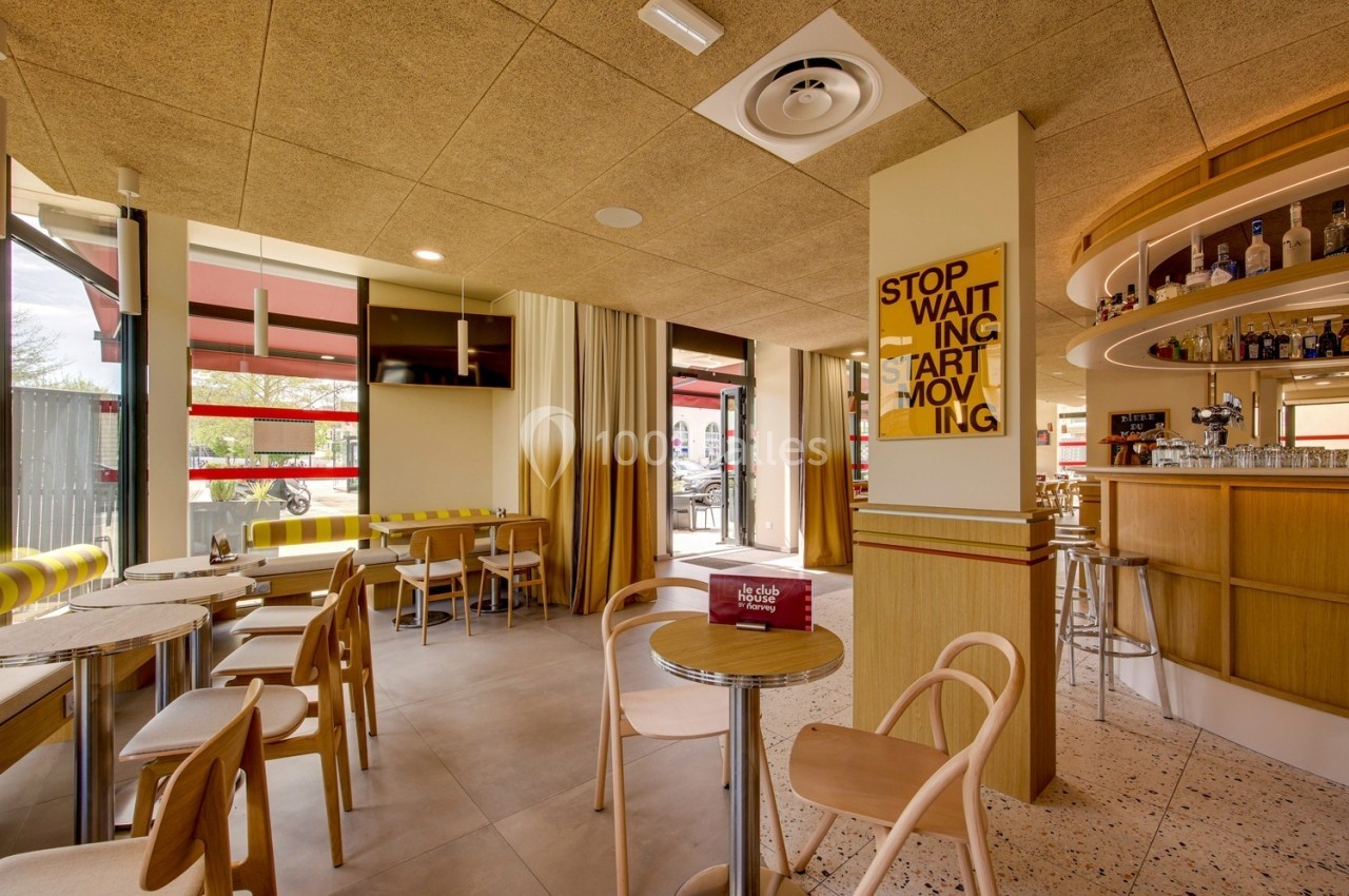 Intérieur d'un café moderne avec tables, chaises en bois, bar équipé et grandes baies vitrées laissant entrer la lumière.