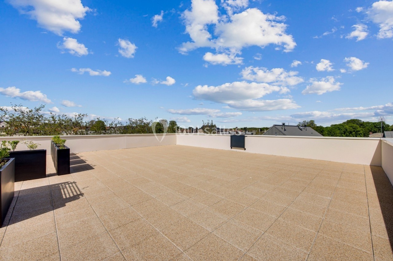 Terrasse spacieuse avec sol en dalles claires, bordée de jardinières, sous un ciel bleu parsemé de nuages.