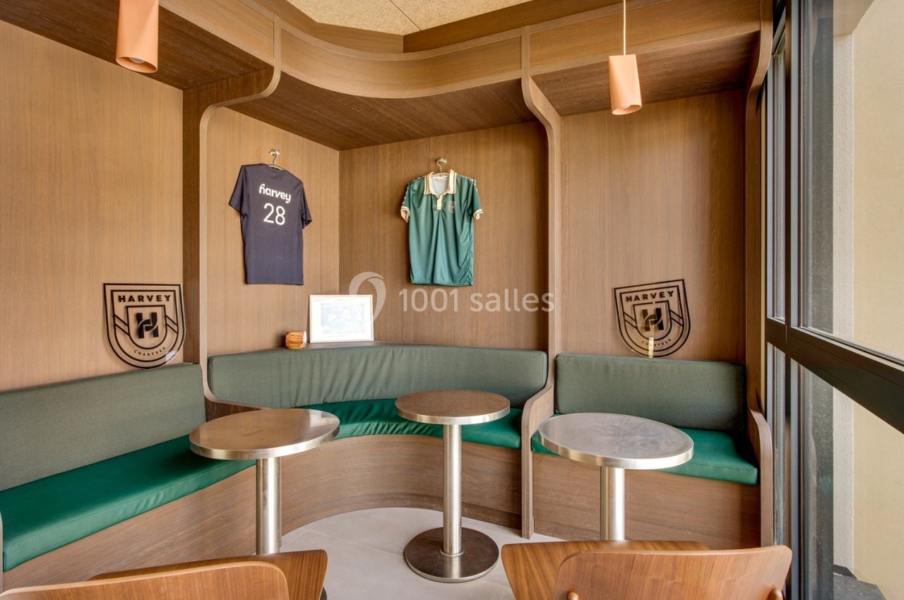 Coin salon avec banquettes vertes, tables rondes et maillots de football accrochés sur des panneaux en bois.