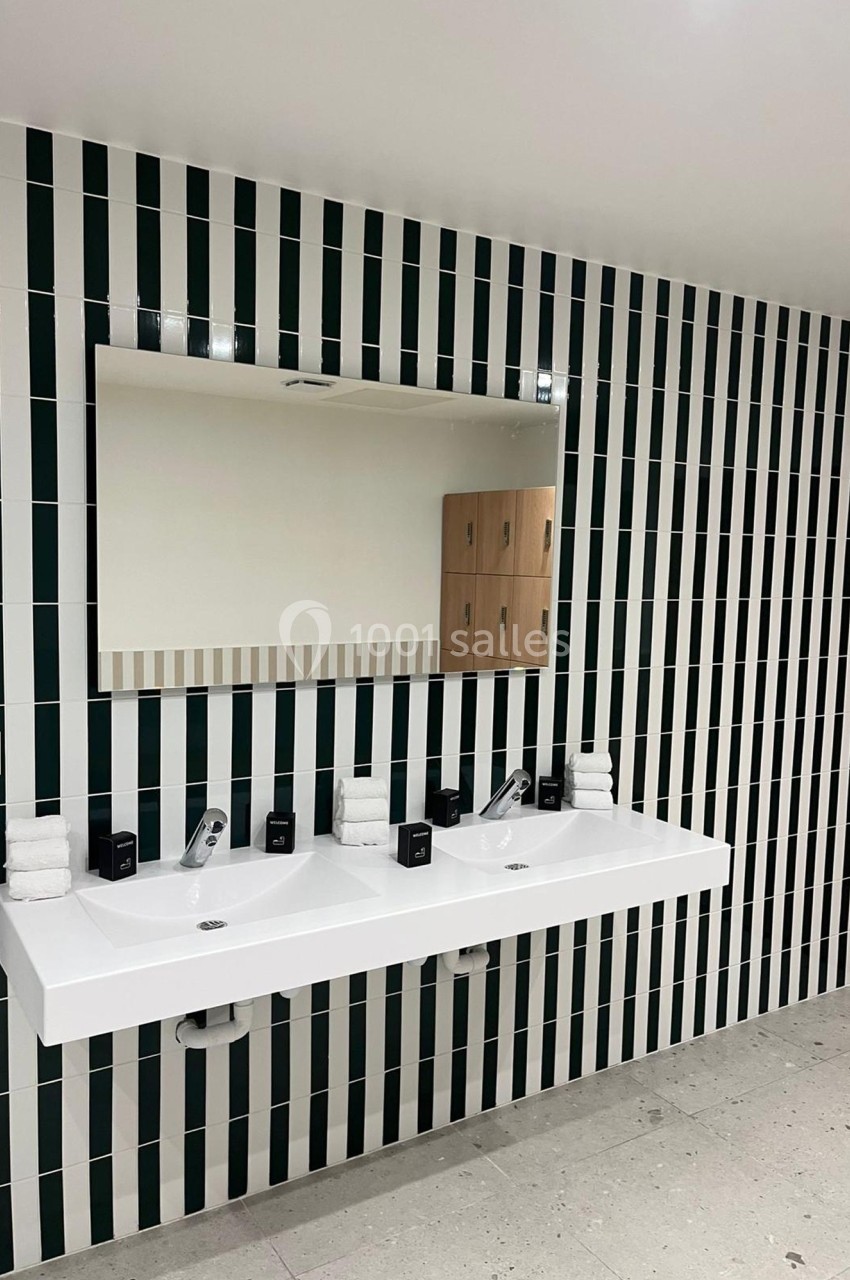 Deux lavabos blancs avec miroir mural, carrelage vert et blanc en arrière-plan, et étagères en bois sur le côté.