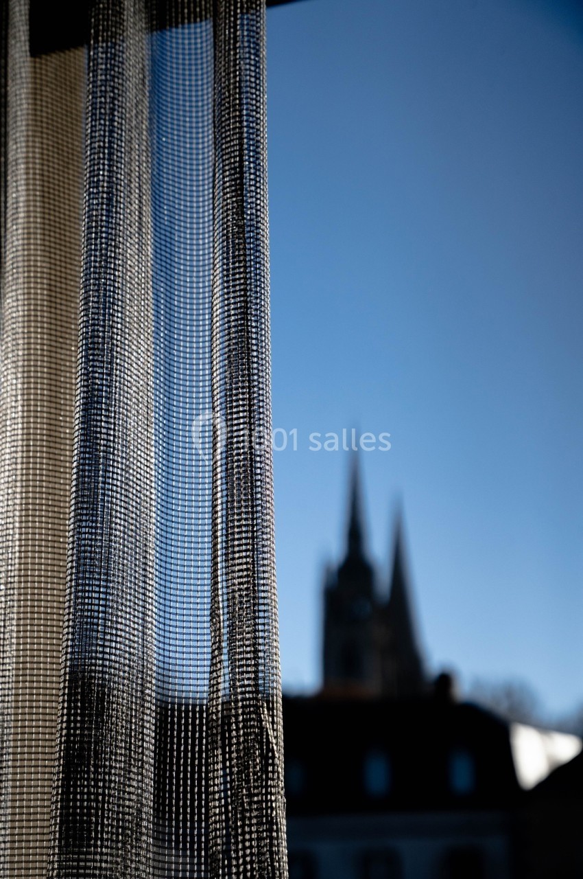 Rideau semi-transparent devant une fenêtre, avec une silhouette floue de bâtiments et un ciel bleu en arrière-plan.