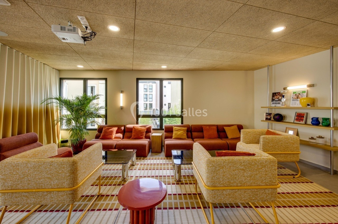 Espace salon lumineux avec fauteuils et canapés colorés, tapis à motifs, étagères décorées et plantes.