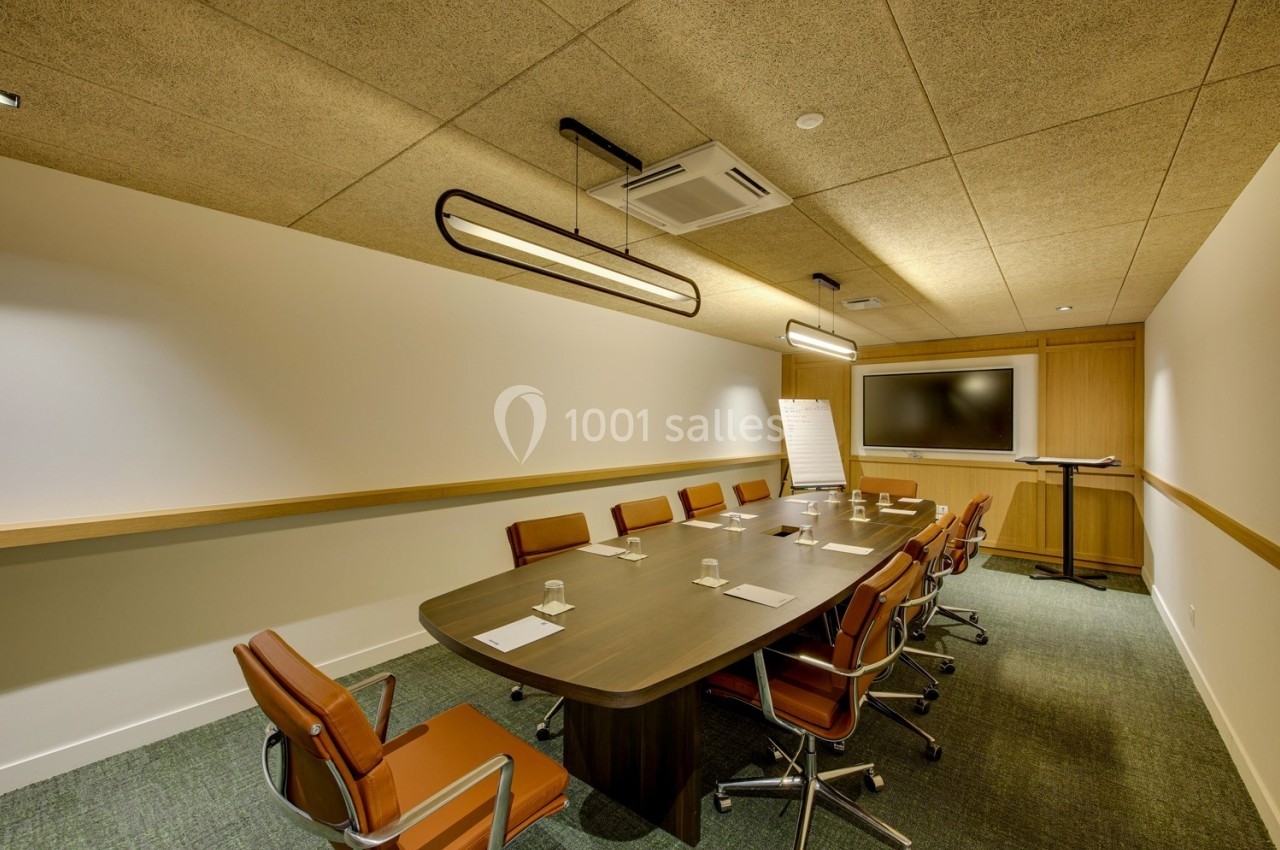 Salle de réunion avec table en bois, chaises en cuir, écran mural, paperboard et éclairage suspendu.