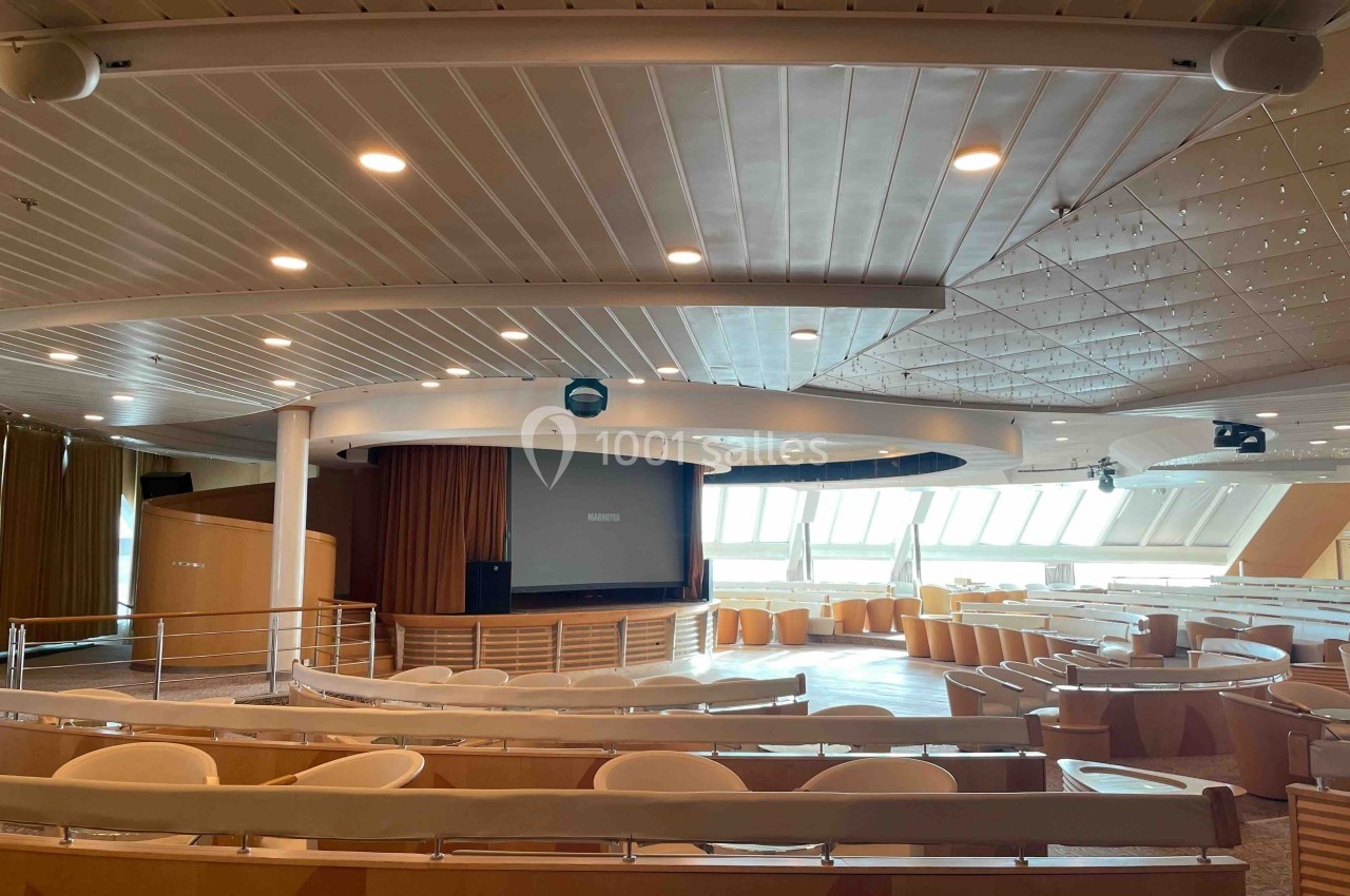 Salle de conférence lumineuse avec sièges disposés en gradins, écran de projection et grandes fenêtres.