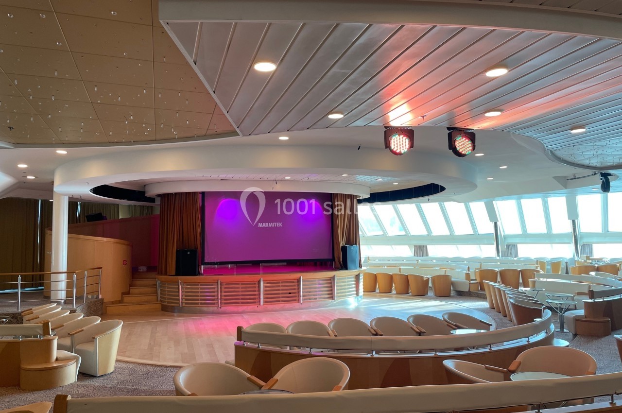 Salle de spectacle moderne avec sièges arrondis, scène centrale, écran projeté et éclairage coloré.