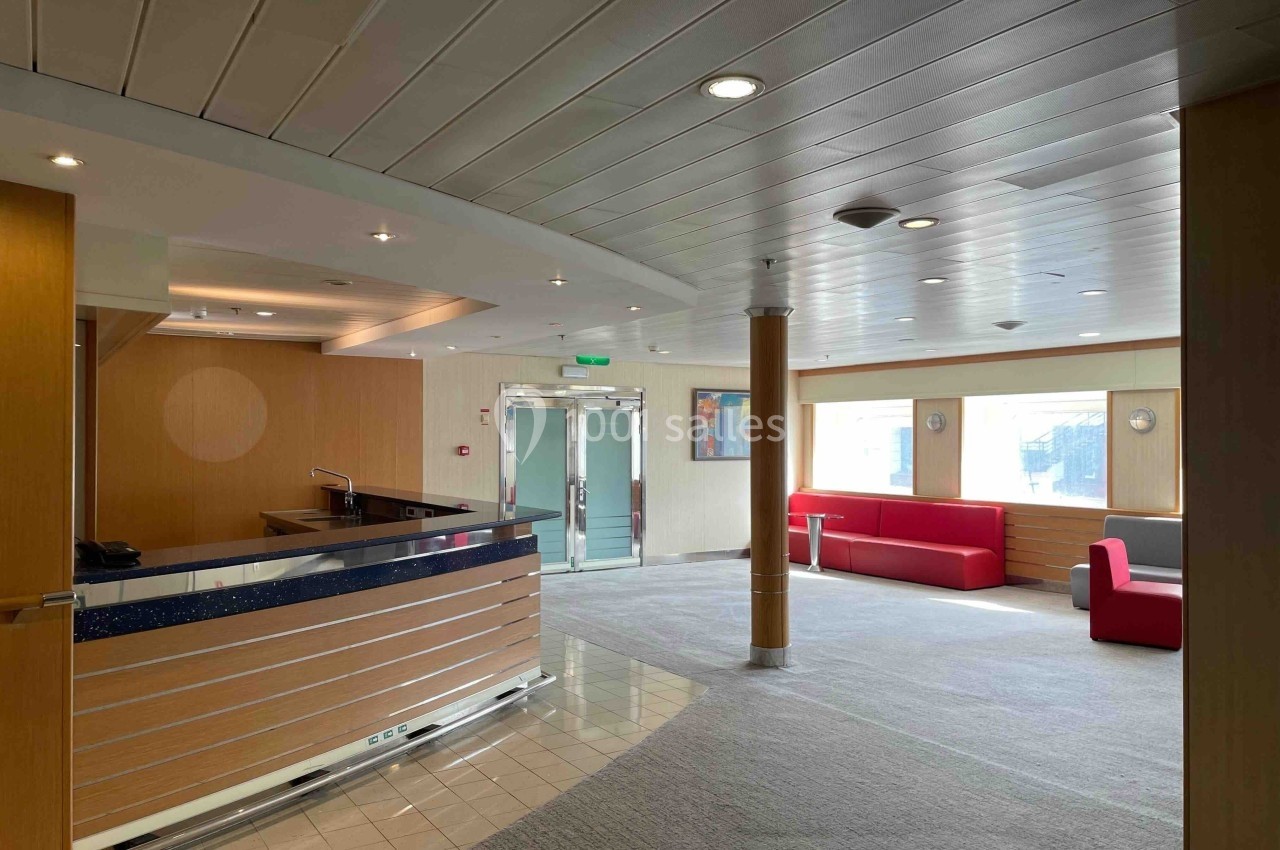 Hall lumineux avec comptoir d'accueil en bois et métal, canapés rouges, grandes fenêtres et éclairage au plafond.