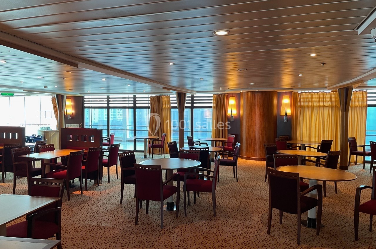 Salle lumineuse avec tables et chaises en bois, grandes fenêtres et rideaux jaunes laissant passer la lumière naturelle.