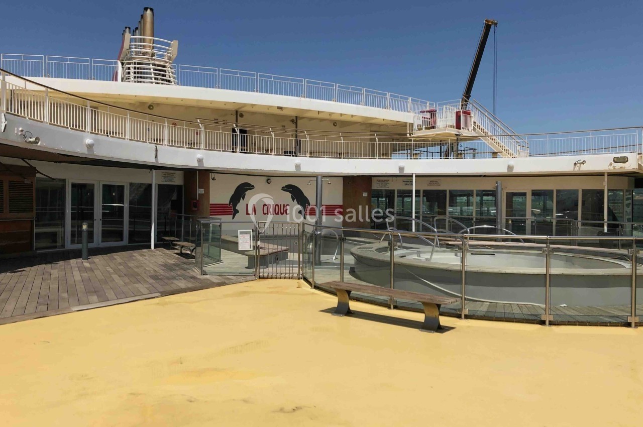 Pont extérieur d'un navire avec sol jaune, piscine vide, banc en bois et mur décoré d'une fresque ’La Cirque’.