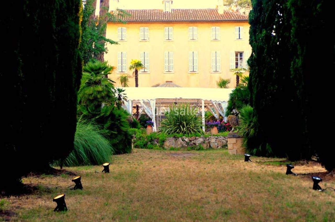 Allée bordée d'arbres menant à une grande maison jaune entourée de végétation et d'un jardin aménagé.