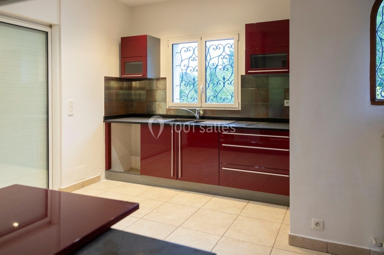 Cuisine moderne avec meubles rouges brillants, plan de travail noir, carrelage beige et fenêtre avec grille décorative.