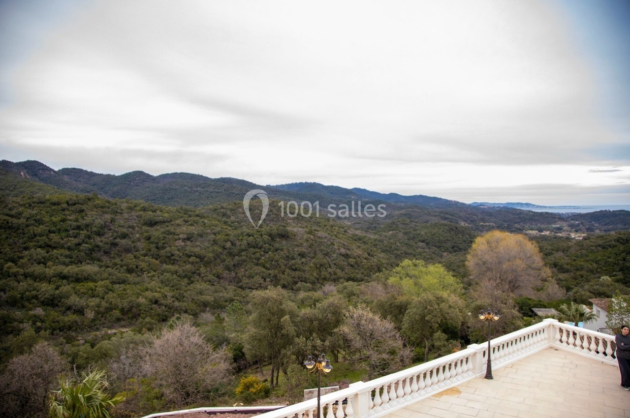 Vue sur un paysage vallonné verdoyant depuis une terrasse avec balustrade blanche, sous un ciel nuageux.