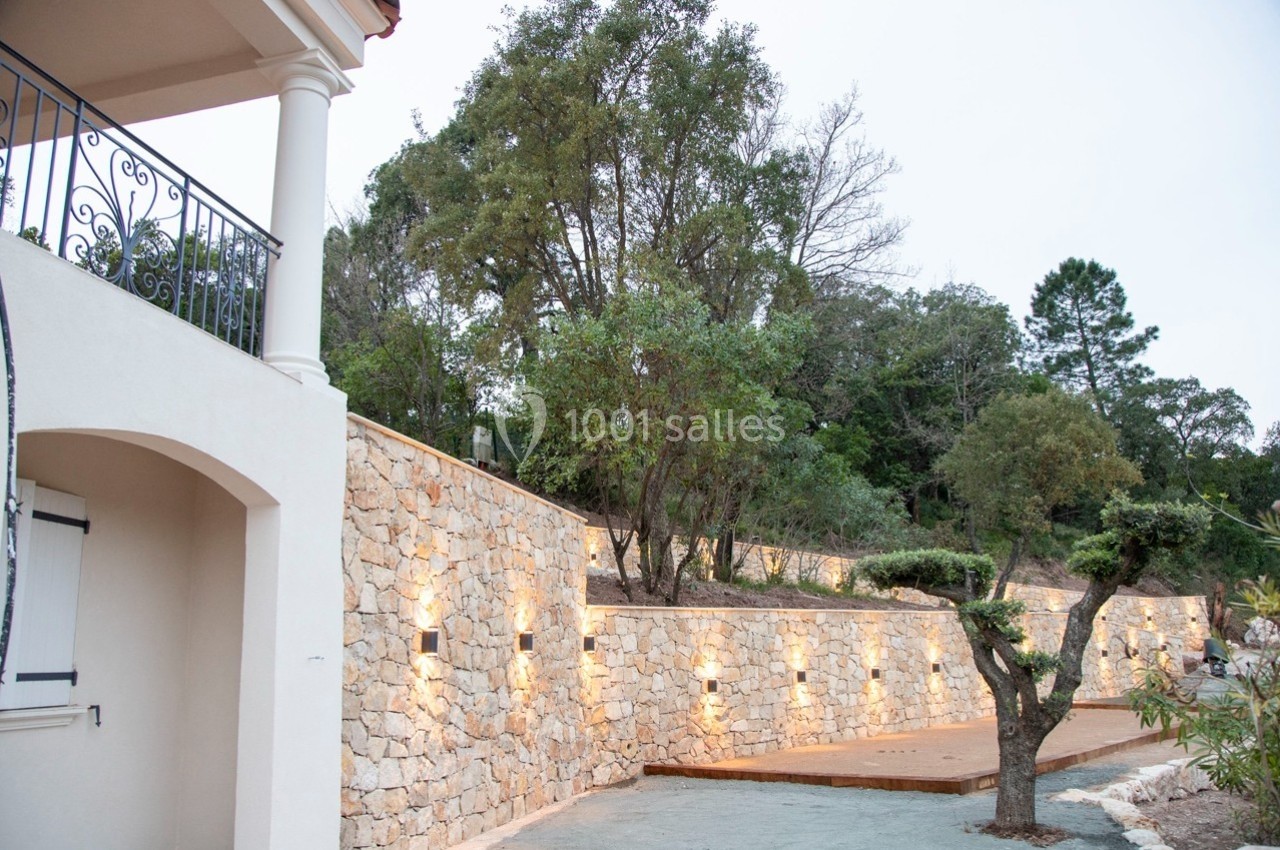 Mur en pierre éclairé par des appliques, bordant un chemin près d'une maison avec balcon, entouré de végétation.