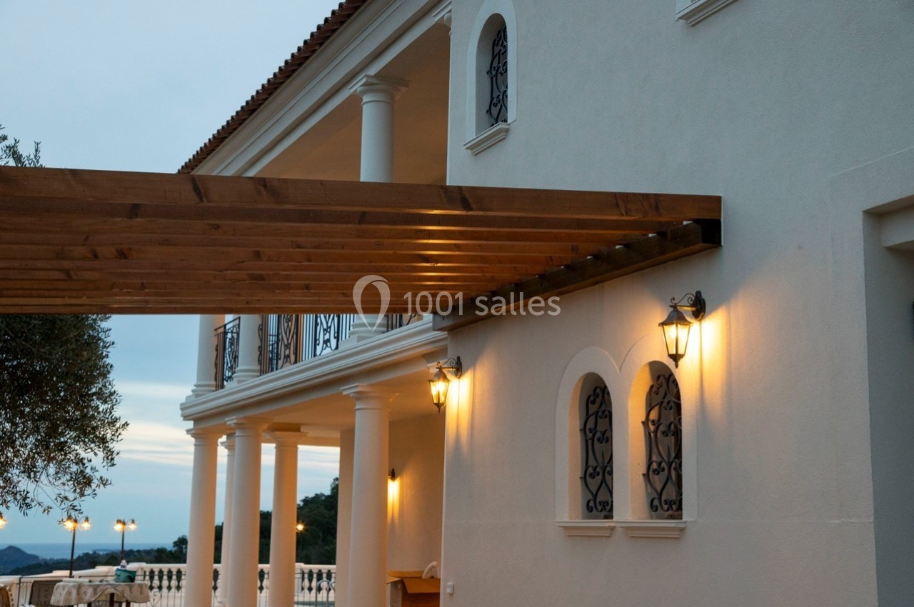 Façade d'une maison avec colonnes blanches, pergola en bois et éclairage extérieur allumé au crépuscule.