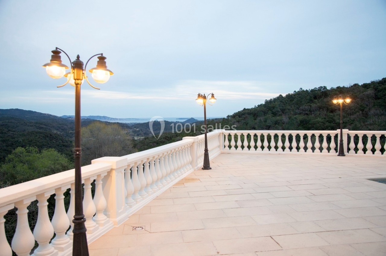 Terrasse en pierre avec balustrade blanche, éclairée par des lampadaires, offrant une vue sur des collines et la mer.