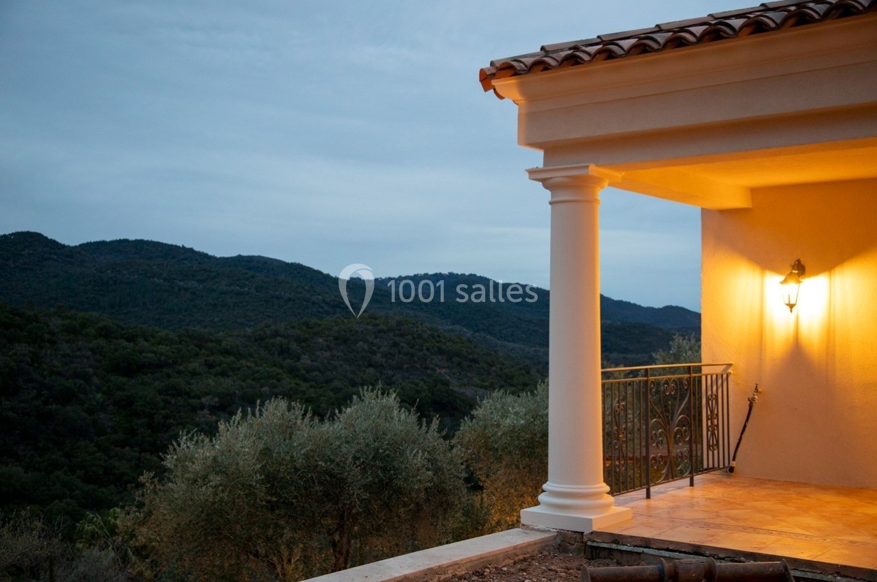 Terrasse éclairée d'une maison avec vue sur des collines boisées sous un ciel nuageux en fin de journée.