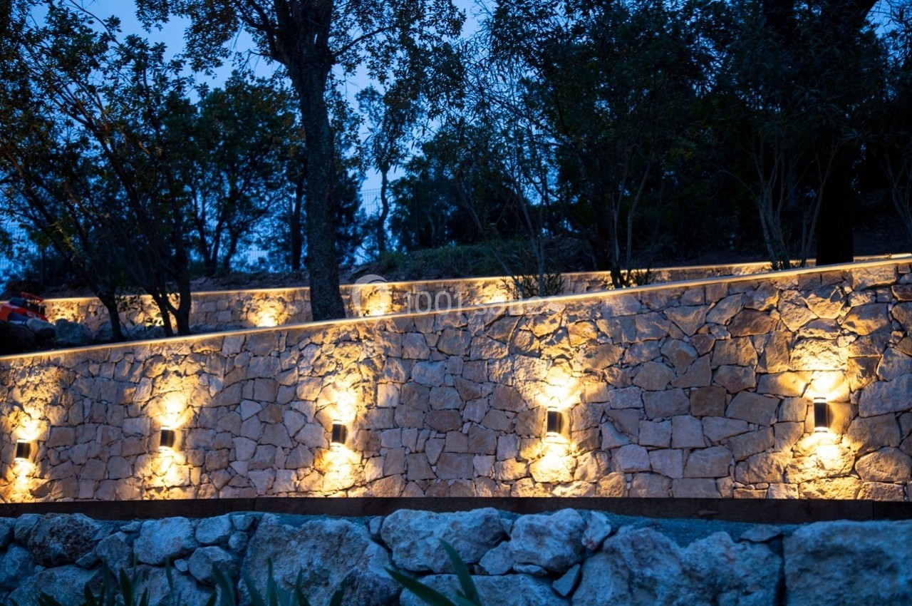 Mur en pierre éclairé par des appliques lumineuses au crépuscule, entouré d'arbres et de végétation.