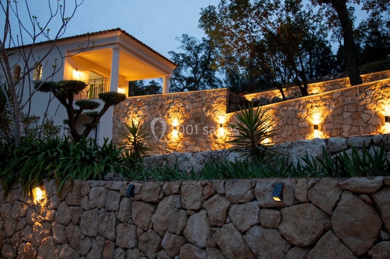 Mur en pierre éclairé par des appliques lumineuses, entouré de végétation, avec une maison en arrière-plan au crépuscule.