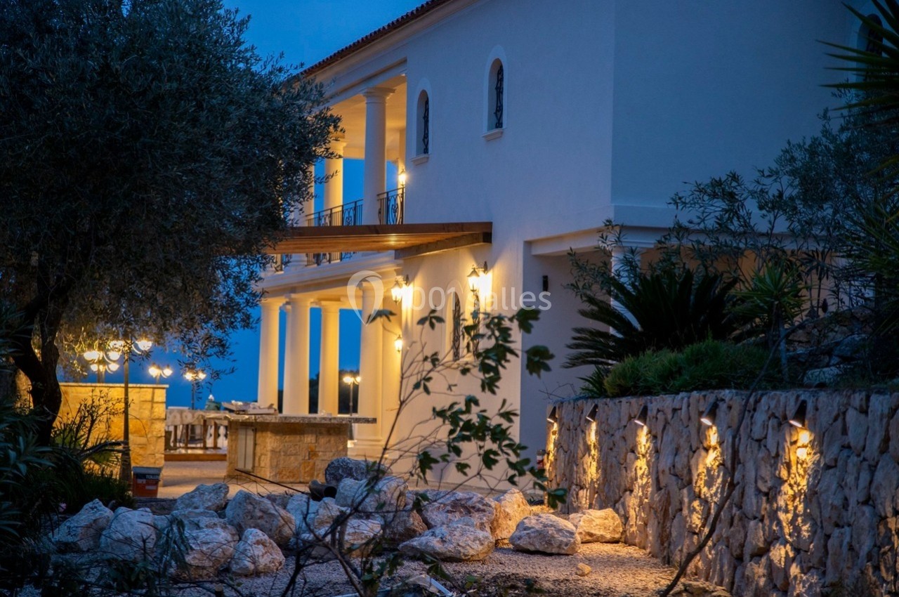 Grande maison blanche éclairée en soirée, entourée de végétation, avec une terrasse et un mur en pierre.