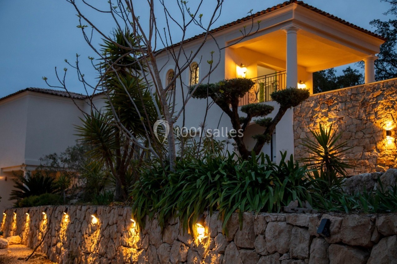 Façade d'une maison méditerranéenne éclairée au crépuscule, entourée de plantes et d'un muret en pierre.