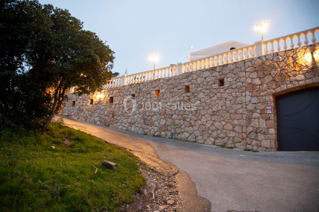 Mur en pierre éclairé par des lampadaires, longeant une route asphaltée bordée de végétation.