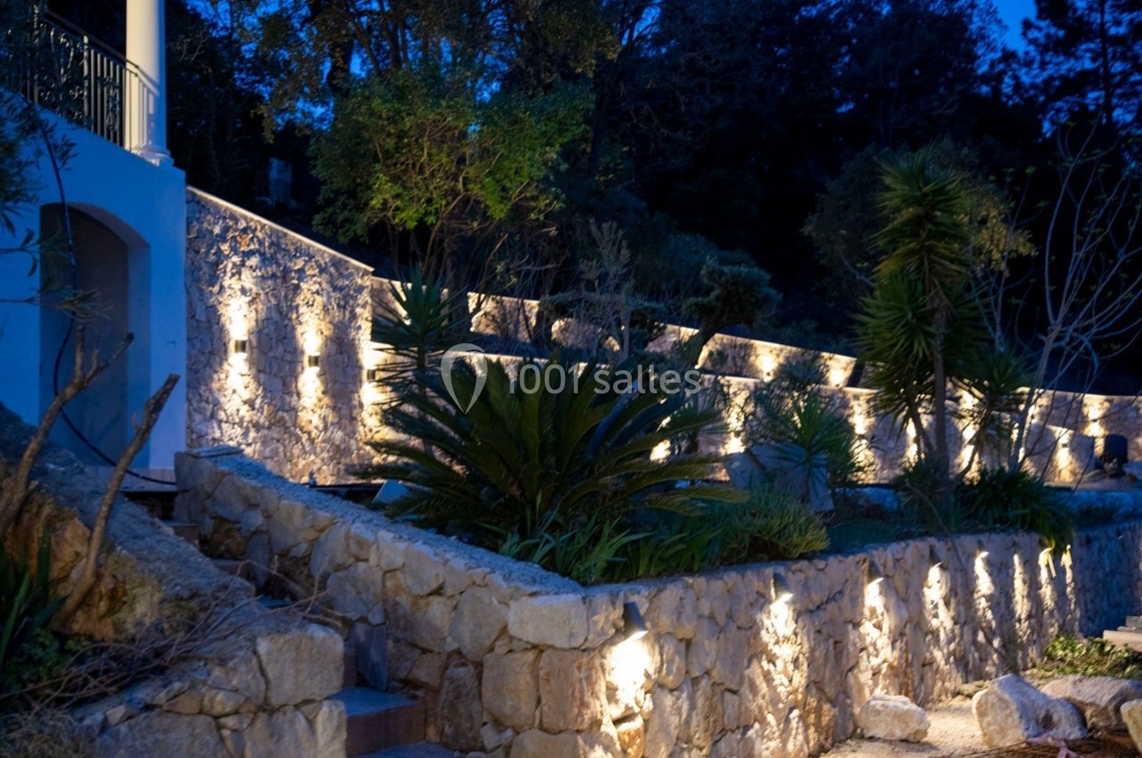 Mur en pierre éclairé par des appliques lumineuses, entouré de végétation dans un jardin au crépuscule.