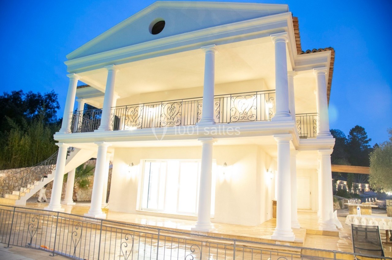 Grande maison blanche à deux étages avec colonnes, éclairée en soirée, entourée de végétation et d'une terrasse.