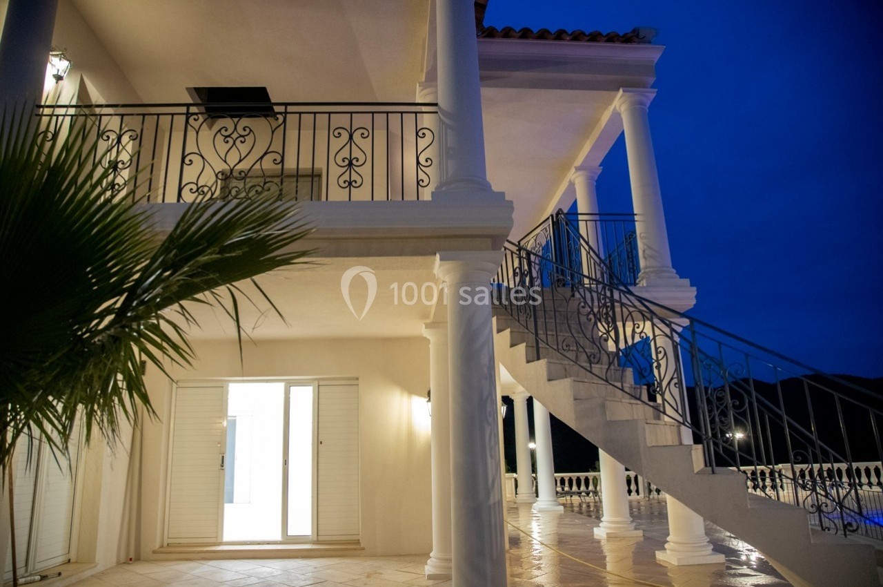 Façade d'une maison avec balcon, escalier extérieur en métal, colonnes blanches et éclairage nocturne.
