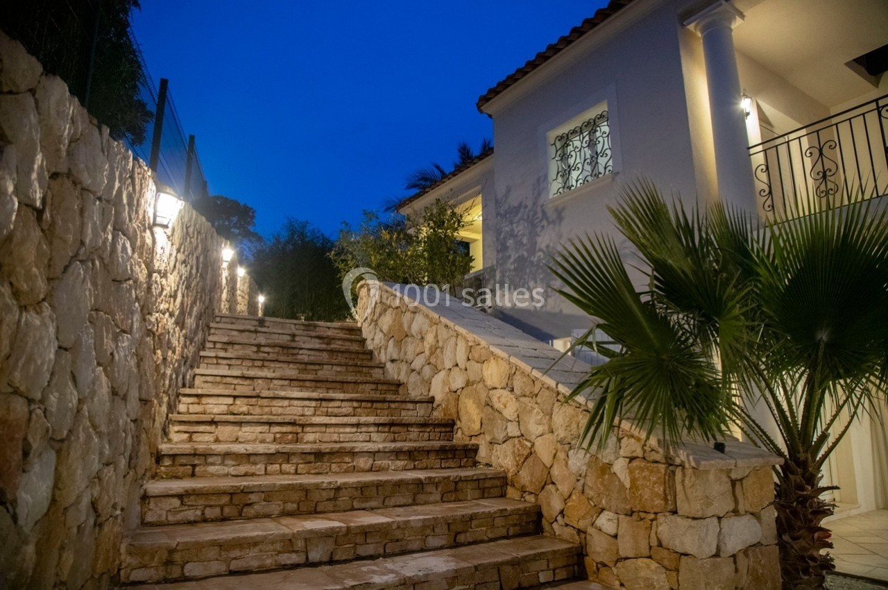 Escalier en pierre éclairé menant à une maison blanche avec des colonnes, entouré de végétation.