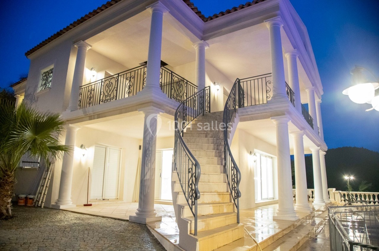 Façade d'une maison luxueuse à deux étages avec colonnes blanches, escalier central et éclairage nocturne.
