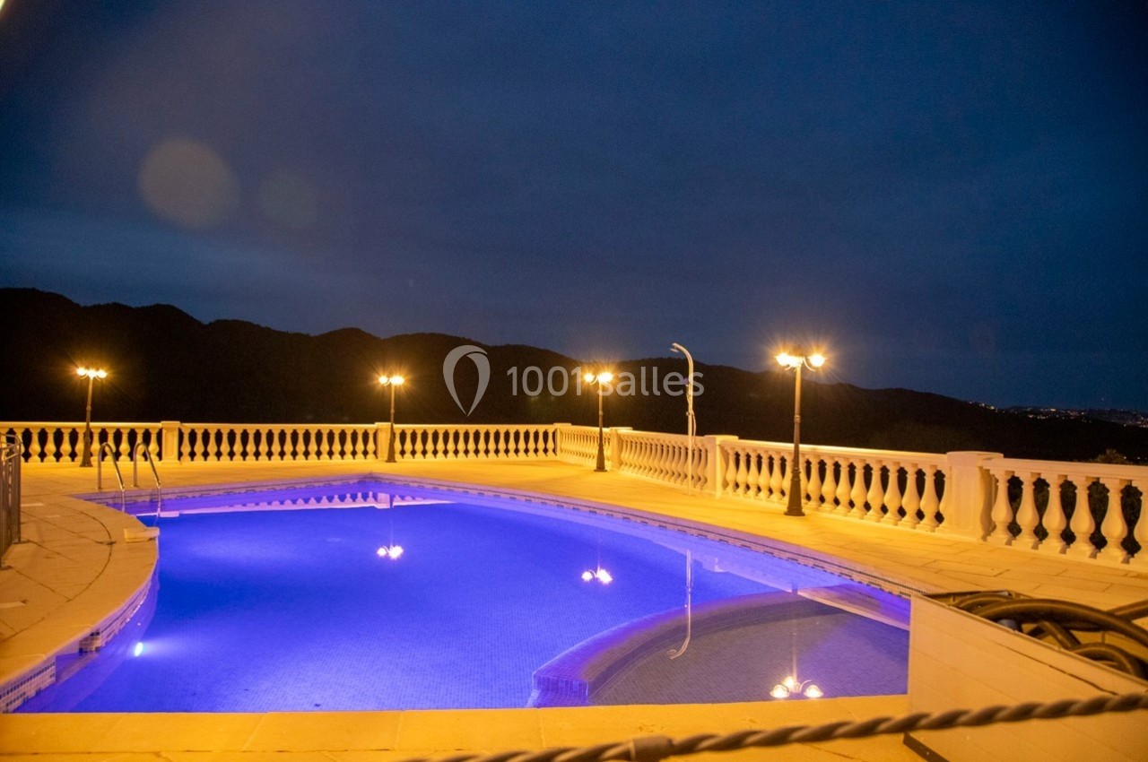 Piscine éclairée en soirée avec balustrades blanches et vue sur des collines sombres à l'arrière-plan.