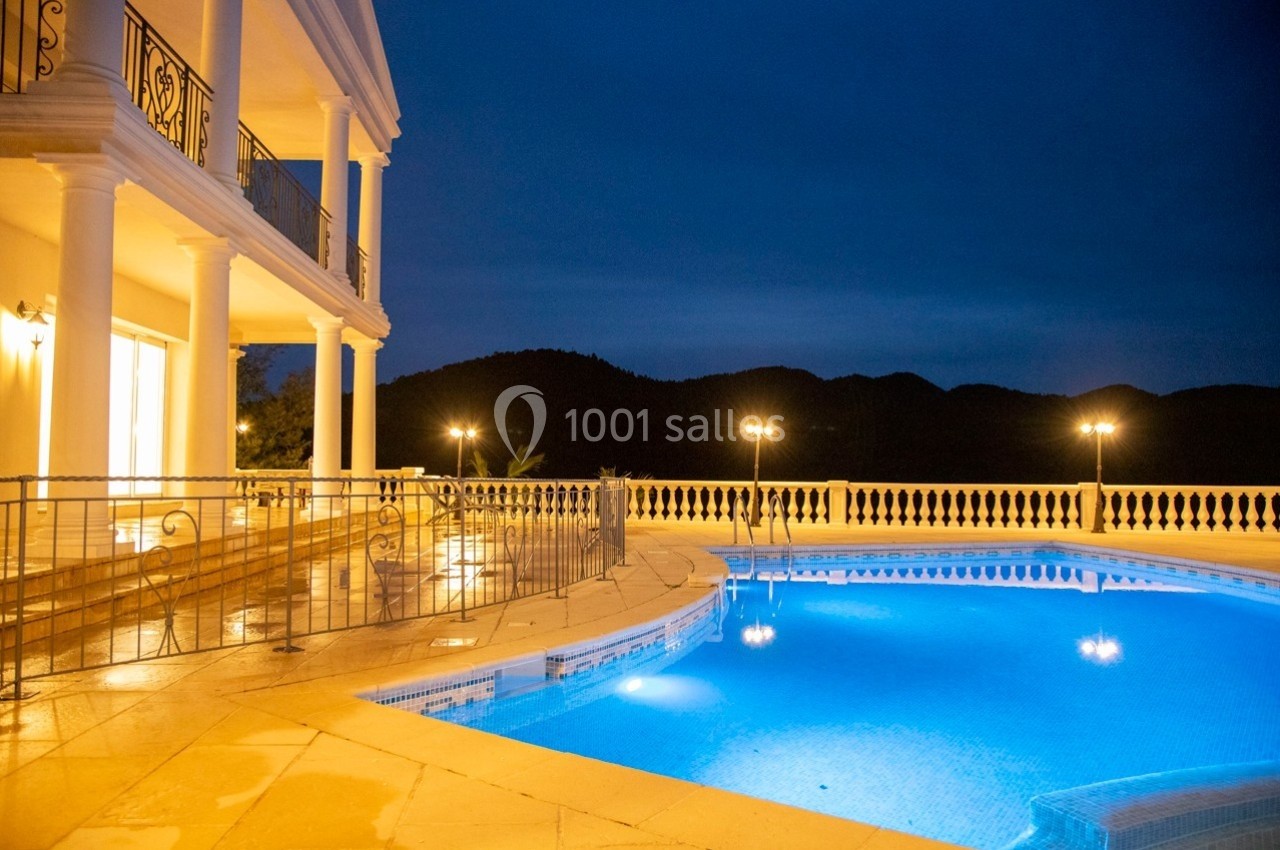 Piscine éclairée entourée d'une terrasse en pierre, devant une villa lumineuse et des collines sombres en arrière-plan.