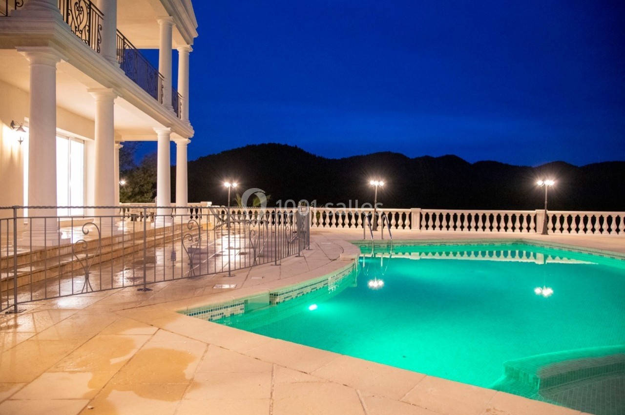 Piscine éclairée de nuit avec terrasse en marbre et balustrades, entourée de collines sombres en arrière-plan.