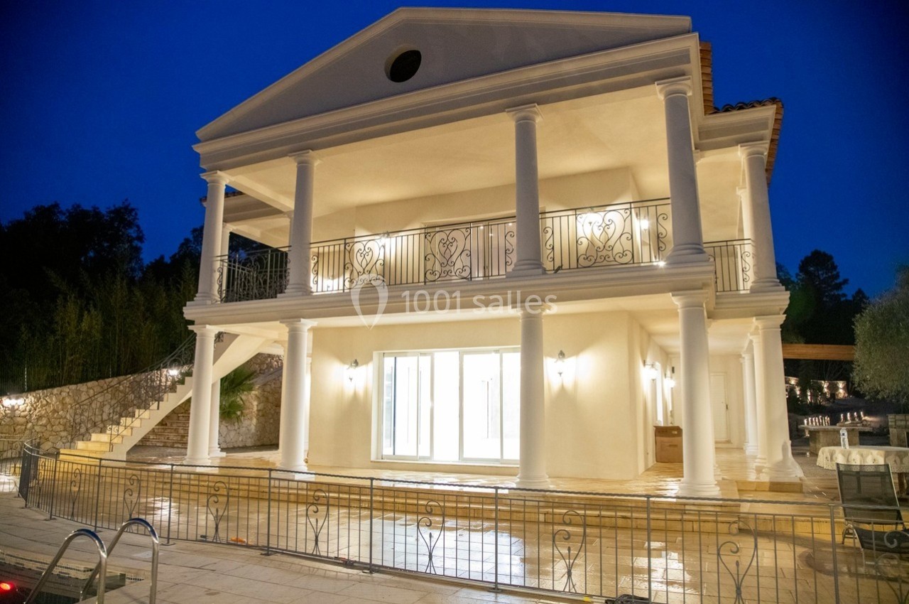 Façade d'une maison de style néoclassique avec colonnes blanches, éclairée la nuit, entourée d'une terrasse et d'une piscine.