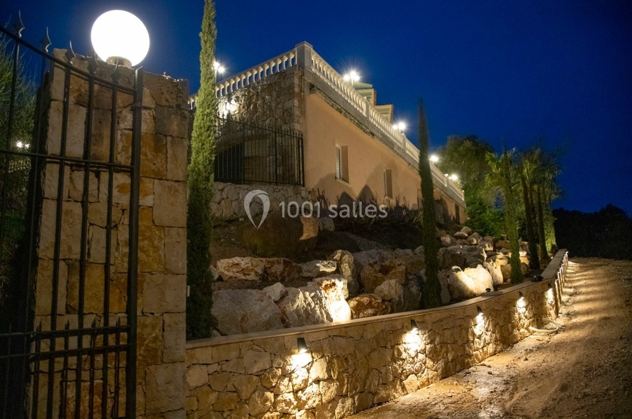 Façade d'une maison en pierre éclairée la nuit, entourée de végétation et d'un chemin bordé de lampes.