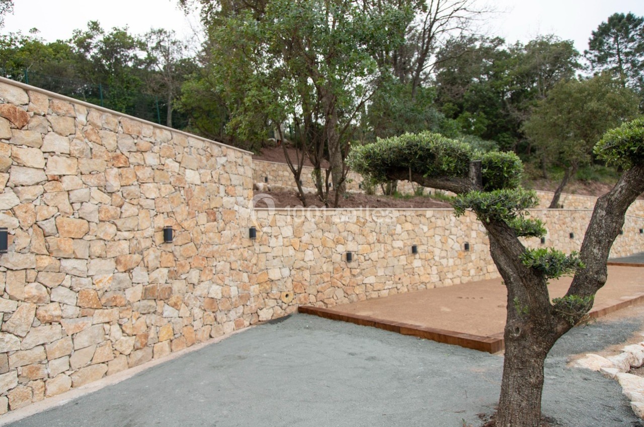 Mur en pierre sèche avec éclairages intégrés, entouré de végétation et d'un sol en gravier et terre battue.