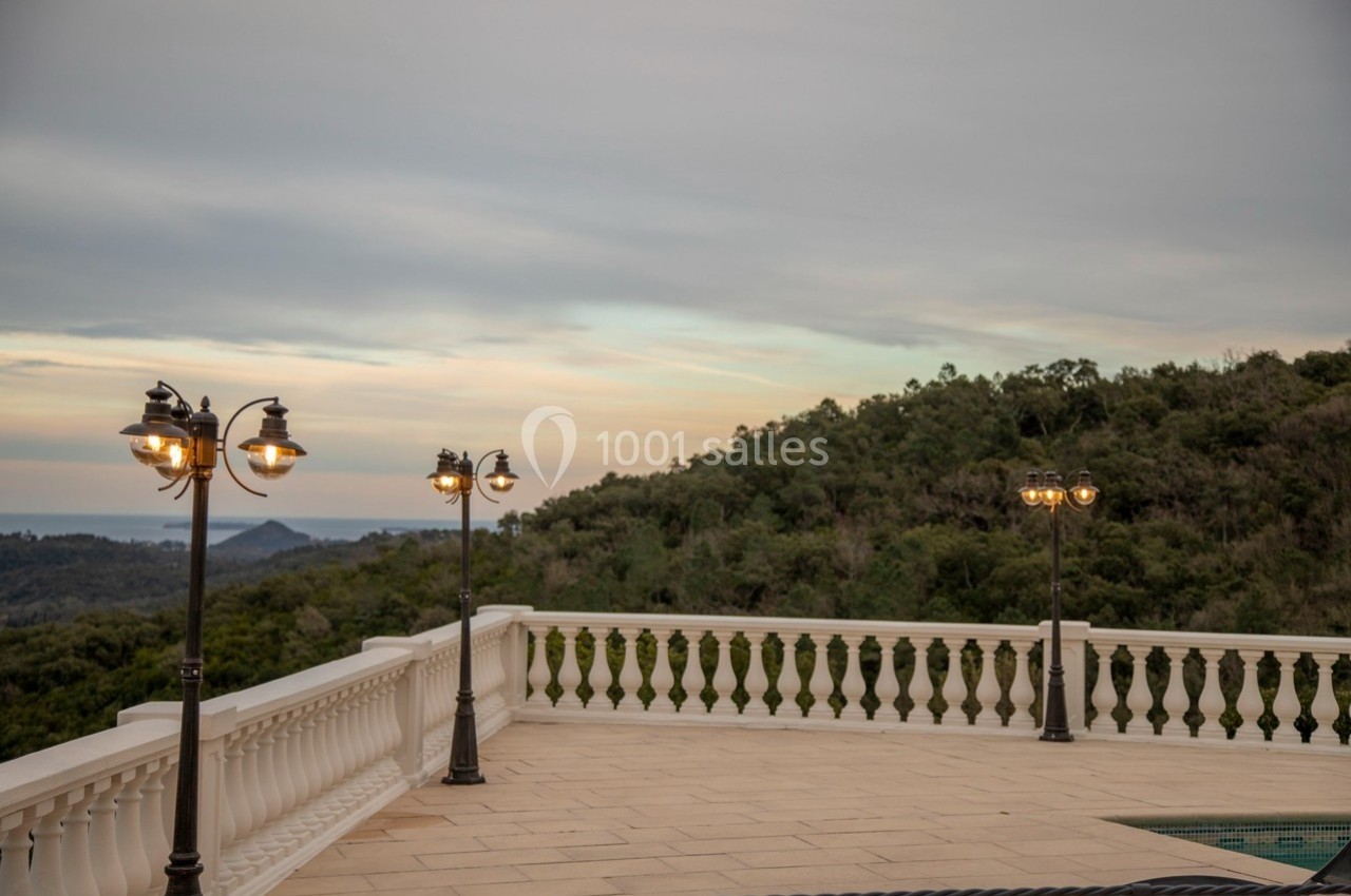 Terrasse avec balustrade, lampadaires allumés et vue sur des collines boisées sous un ciel nuageux.
