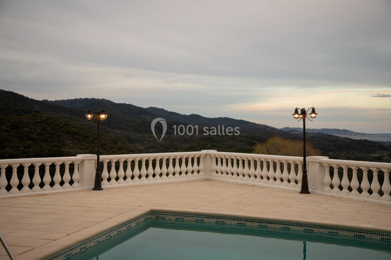 Terrasse avec piscine, balustrade blanche, lampadaires allumés et vue sur des collines sous un ciel nuageux.
