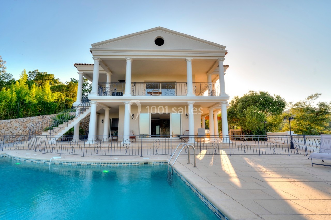 Grande maison blanche à deux étages avec colonnes, terrasse et piscine au premier plan, entourée de verdure.