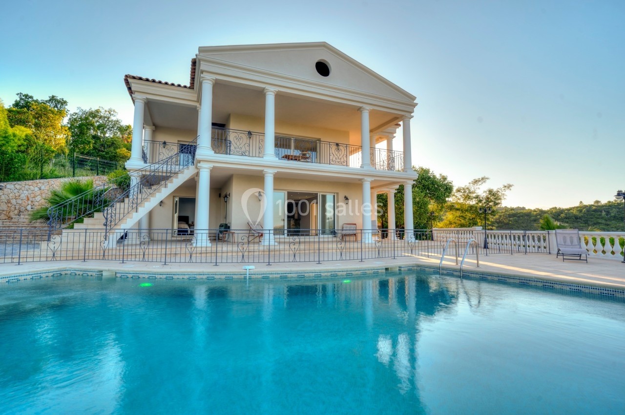 Villa à deux étages avec colonnes blanches, terrasse et piscine, entourée de verdure sous un ciel dégagé.