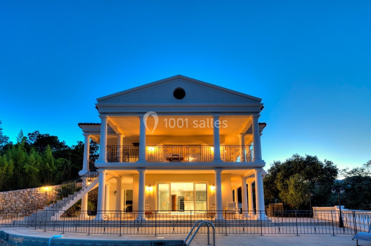 Façade d'une grande maison à deux étages avec colonnes, éclairée la nuit, entourée de végétation.
