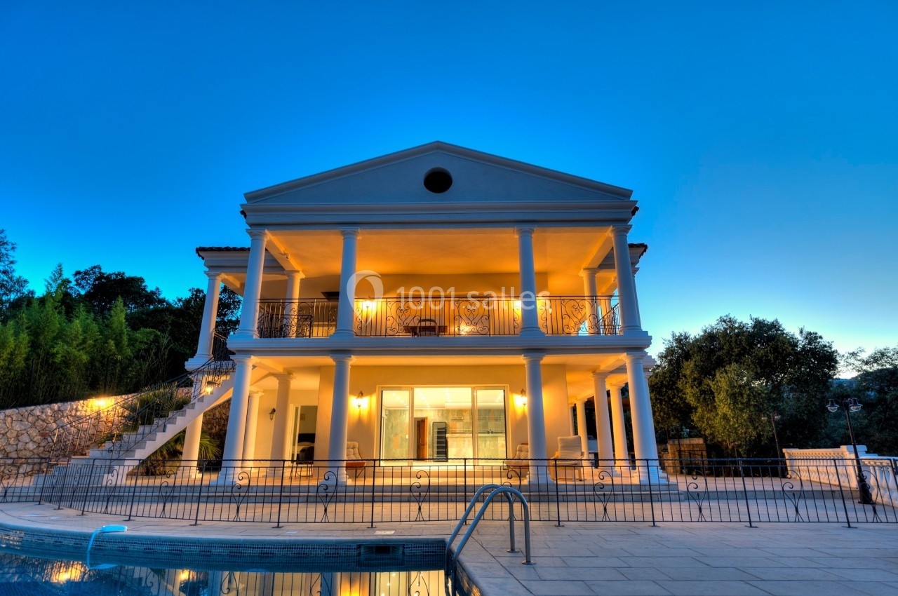 Façade d'une grande maison avec colonnes blanches, éclairée au crépuscule, devant une piscine et un jardin.