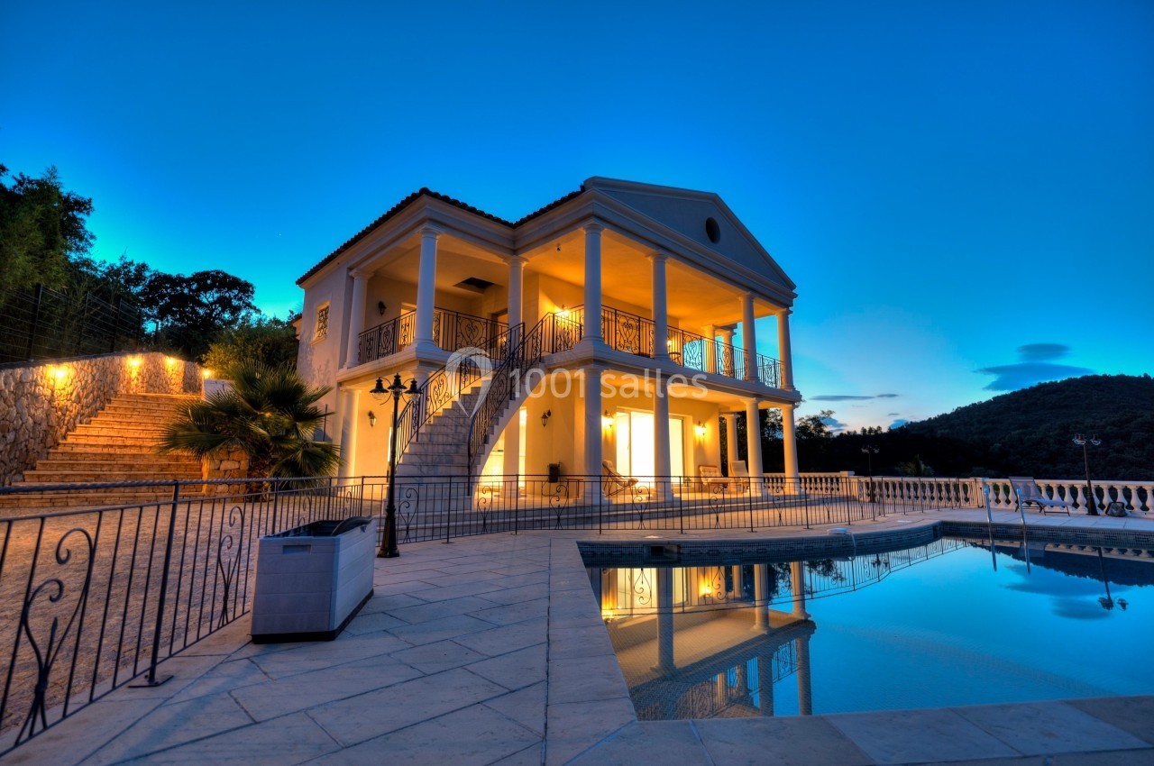 Villa élégante avec colonnes, éclairée au crépuscule, entourée d'une terrasse et d'une piscine reflétant le ciel.